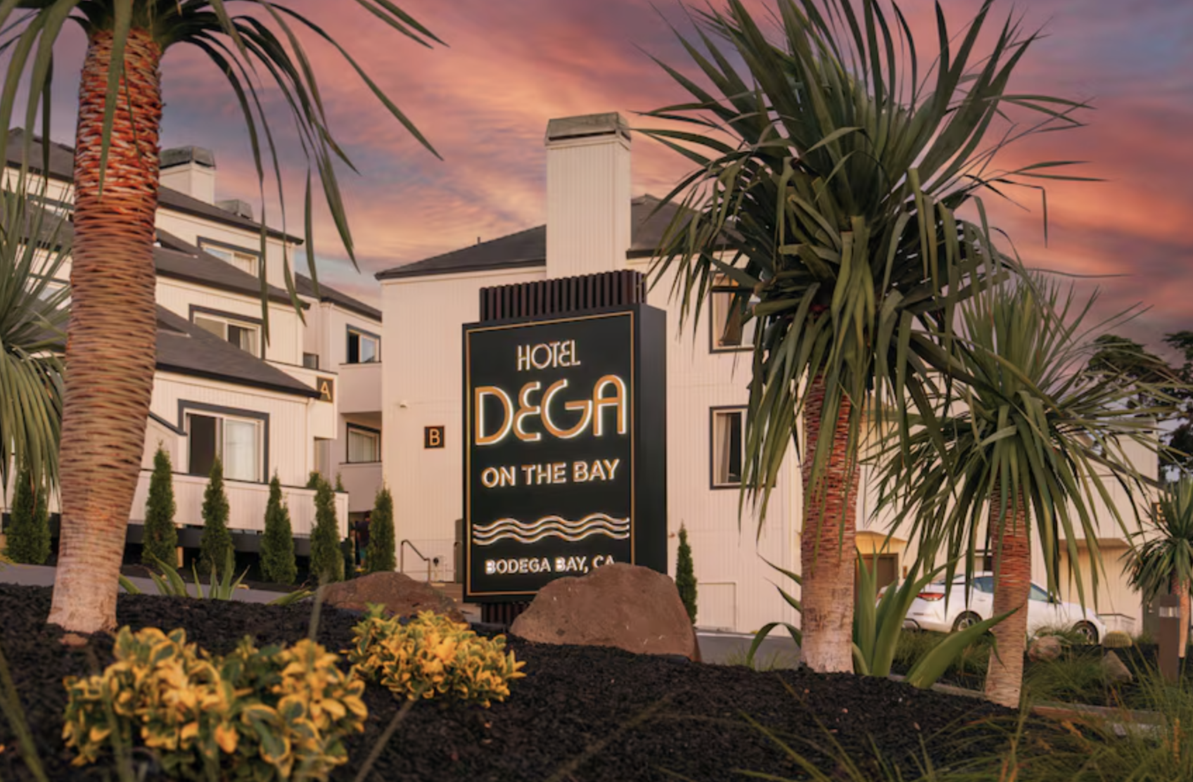 Exterior view of Hotel Deja on the Bay in Bodega Bay, California, during sunset with pink clouds, palm trees, and a sign in the foreground.