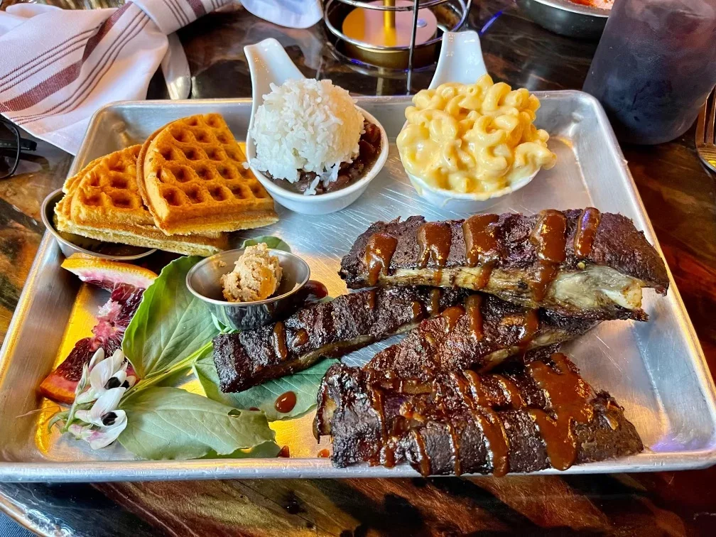 A metal tray with ribs topped with barbecue sauce, served with baked waffle pieces, vanilla ice cream, macaroni and cheese, a small bowl of a paste or sauce, and some garnished fruit and flowers.