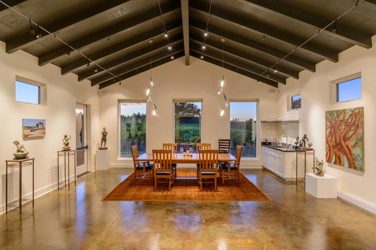 Interior view of a dining room with a wooden table and chairs on an orange rug, art and sculptures on the walls and pedestals, large windows showing an outdoor landscape, and a built-in bar area to the right, with a vaulted ceiling and track lighting.
