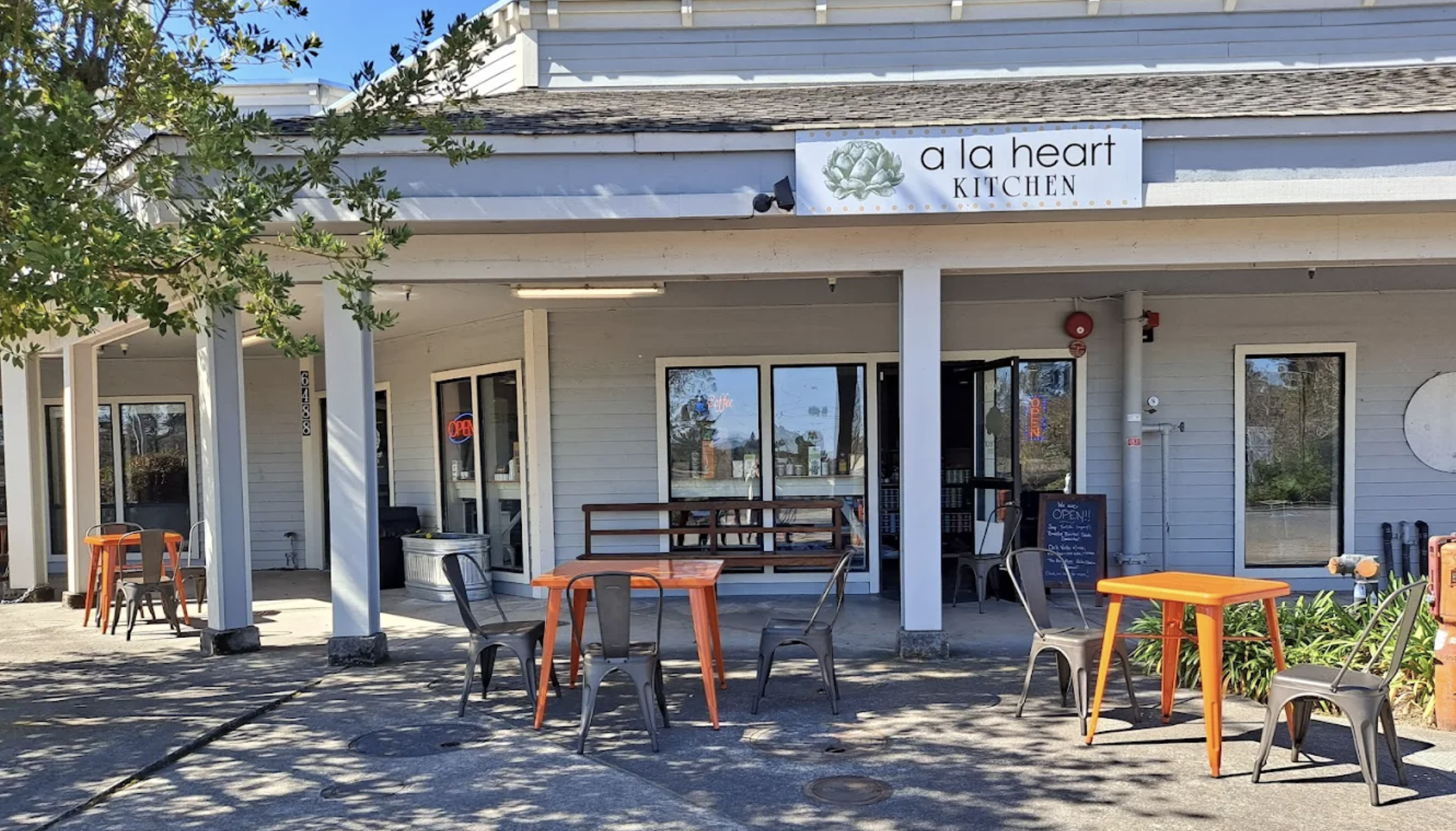 Exterior view of a restaurant named 'a la heart Kitchen' with outdoor seating, including orange tables and metal chairs, and a glass door with neon 'Open' sign.