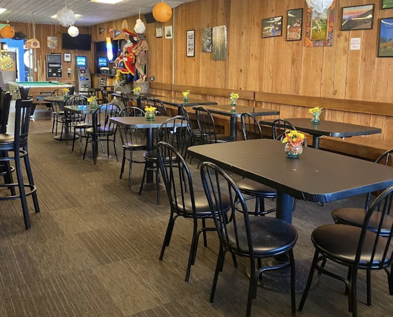 Empty restaurant with black tables and chairs, yellow flowers in vases, wooden wall with framed pictures, pool table, and arcade machines in the background.