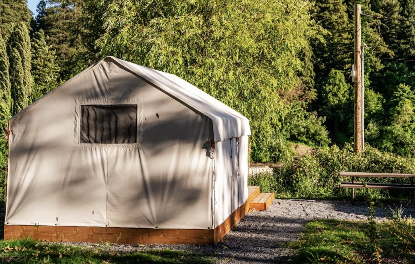 A white canvas tent on a wooden platform in a lush green outdoor setting with trees, shrubs, and a wooden bench.