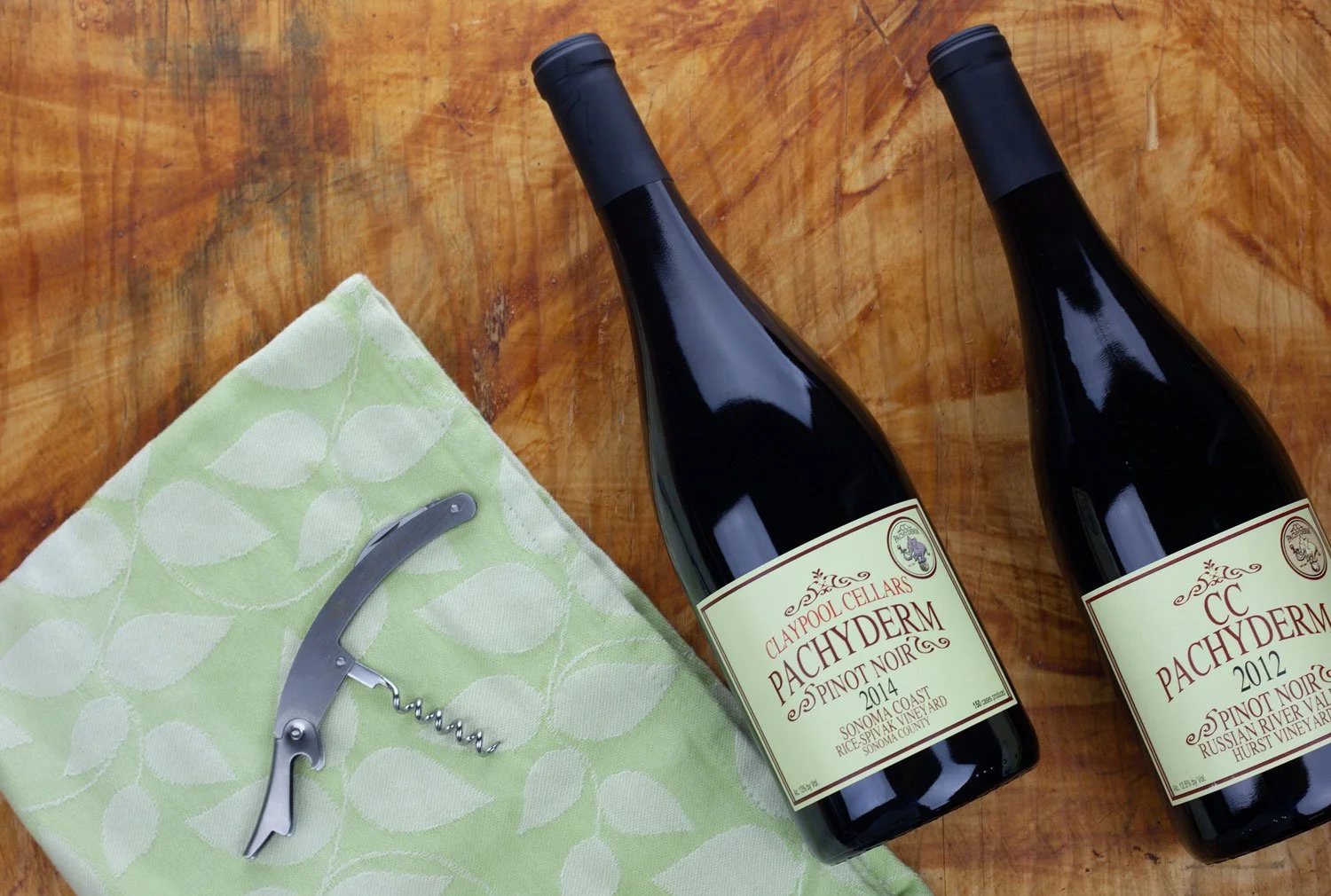 Two bottles of Pachyderm Pinot Noir wine on a wooden table, with a green cloth and a silver corkscrew on the left.