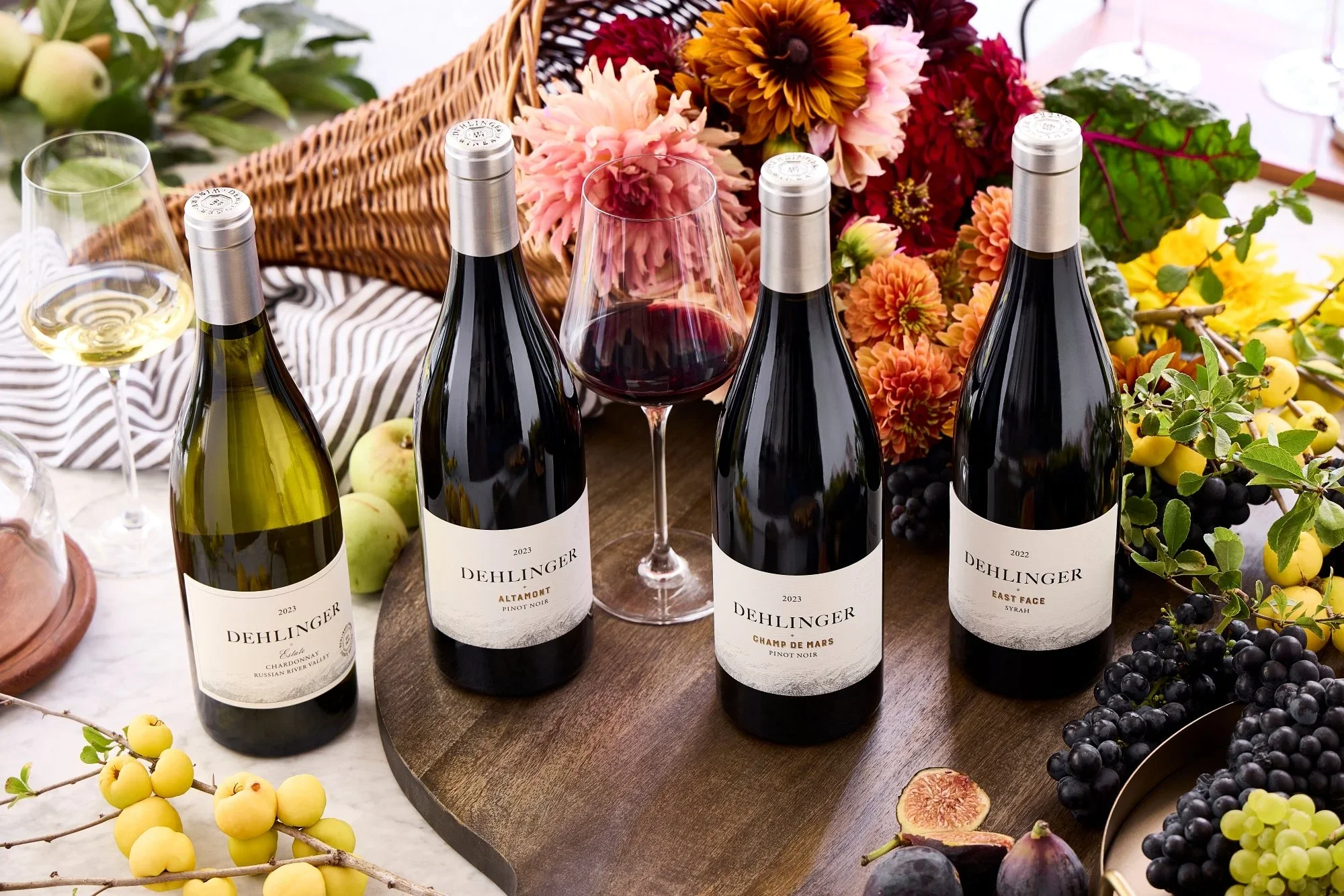 Four bottles of Dehlinger wine on a wooden table, with a glass of white wine and a glass of red wine, surrounded by grapes, figs, apples, and colorful flowers.