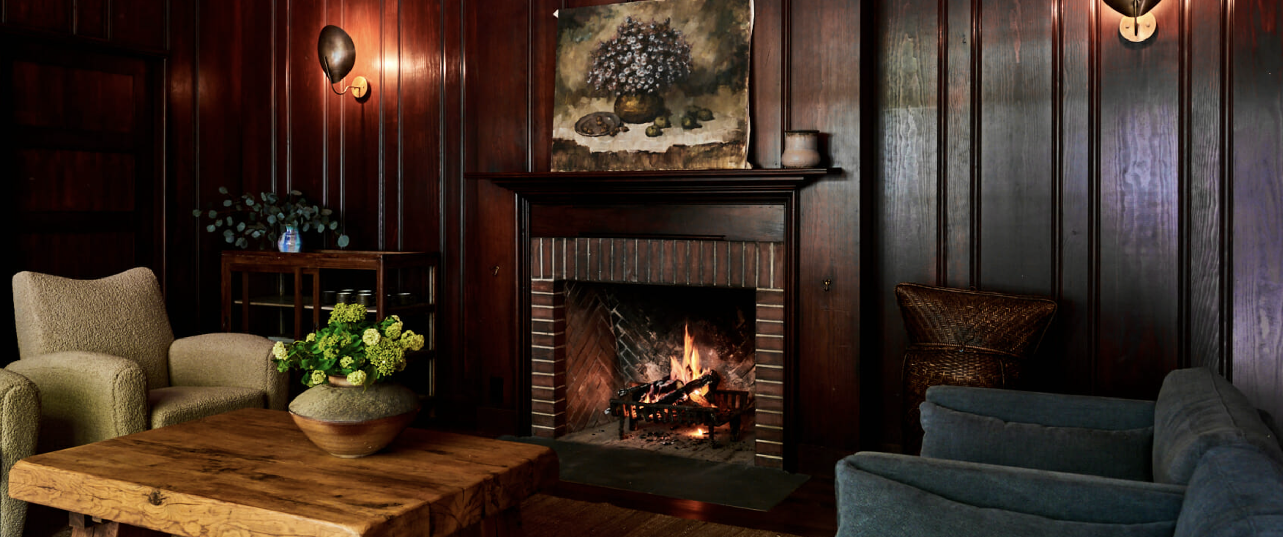 Living room with dark wood-paneled walls, fireplace with a burning fire, beige armchair, blue sofa, wooden coffee table with a green floral centerpiece, artwork above the fireplace, wall sconces lighting the room.