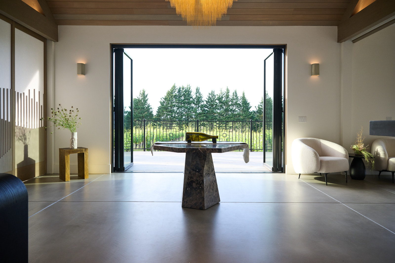 Modern living room with open sliding doors leading to a balcony overlooking a lush green landscape with trees, a marble table in the center, and contemporary furniture including chairs and vases.
