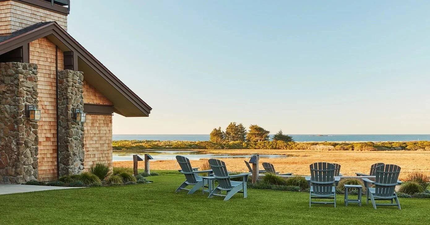 Adirondack chairs on a grassy yard near water with trees and ocean in the background, next to a house with stone and shingle exterior.