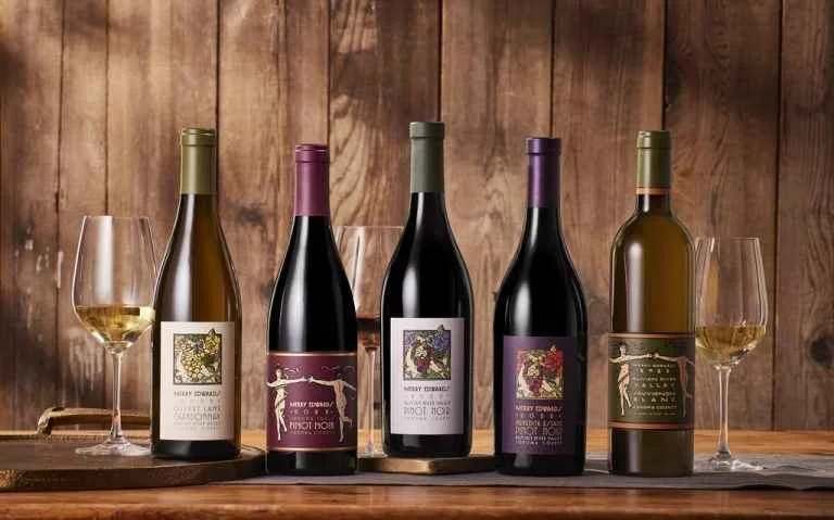 Five bottles of wine with four glasses of white wine, set on a wooden table against a wood-paneled wall.
