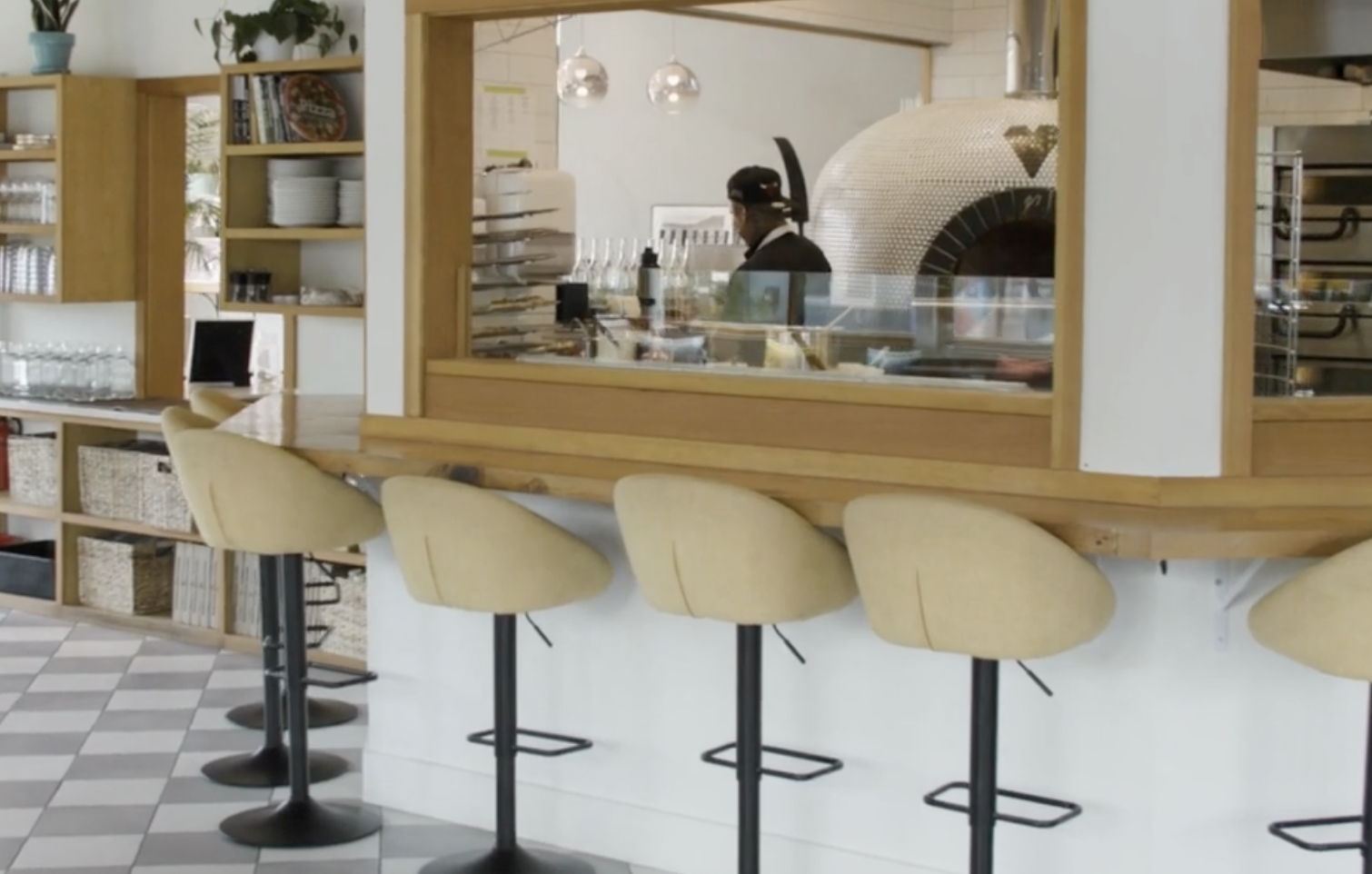 View of a modern cafe or restaurant bar area with four yellow bar stools, a wooden counter, and a chef preparing food behind a glass partition with a pizza oven in the background.