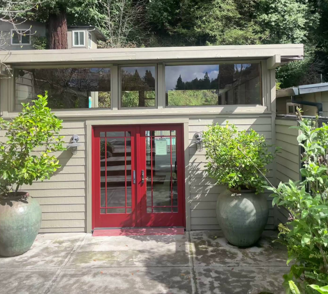 A building with a red double door entrance and large potted green plants on either side. The building has beige siding and a row of windows above the door, with trees visible in the background.