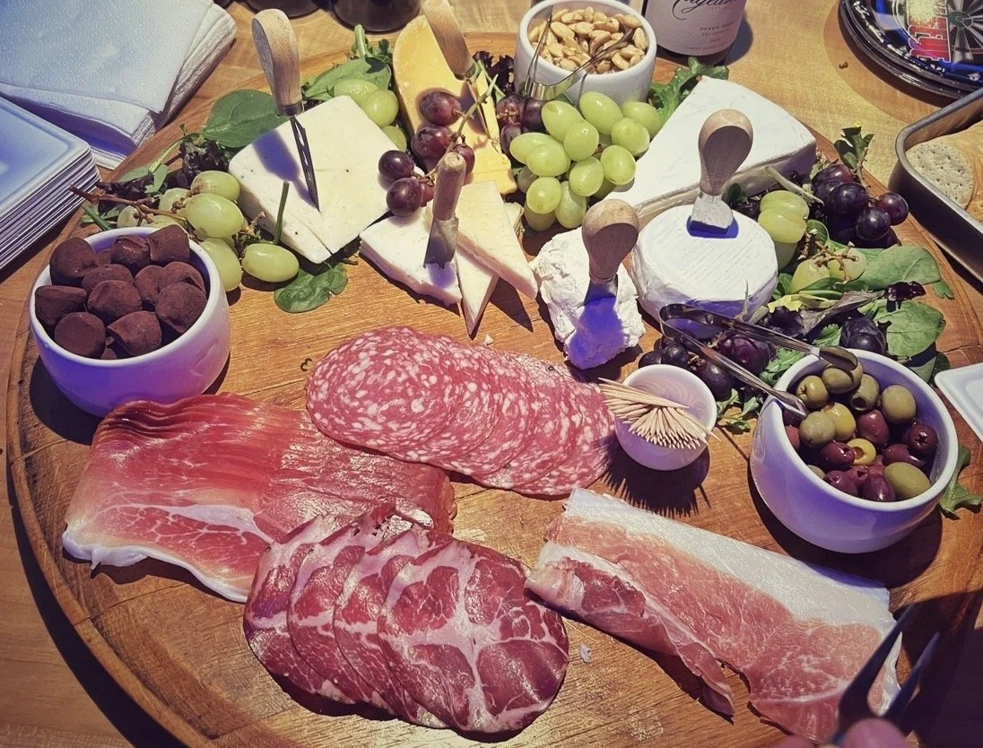 Cheese, grapes, nuts, cured meats, olives, and bread on a wooden table, suggesting a cheese board or platter.