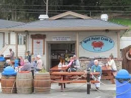 People gathered outside a seafood restaurant called 'Wild Point Crab Co' with a sign featuring a crab logo, outdoor seating, and large barrels used as tables.