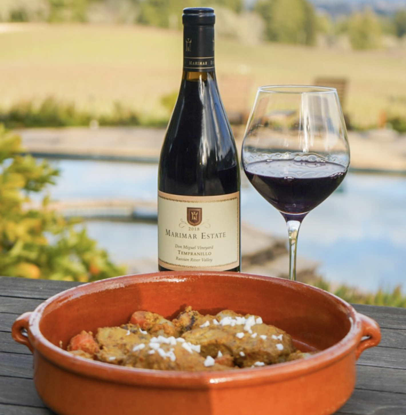 A bottle of red wine, a glass of red wine, and a dish of cooked food with vegetables in an outdoor setting with a river and trees in the background.