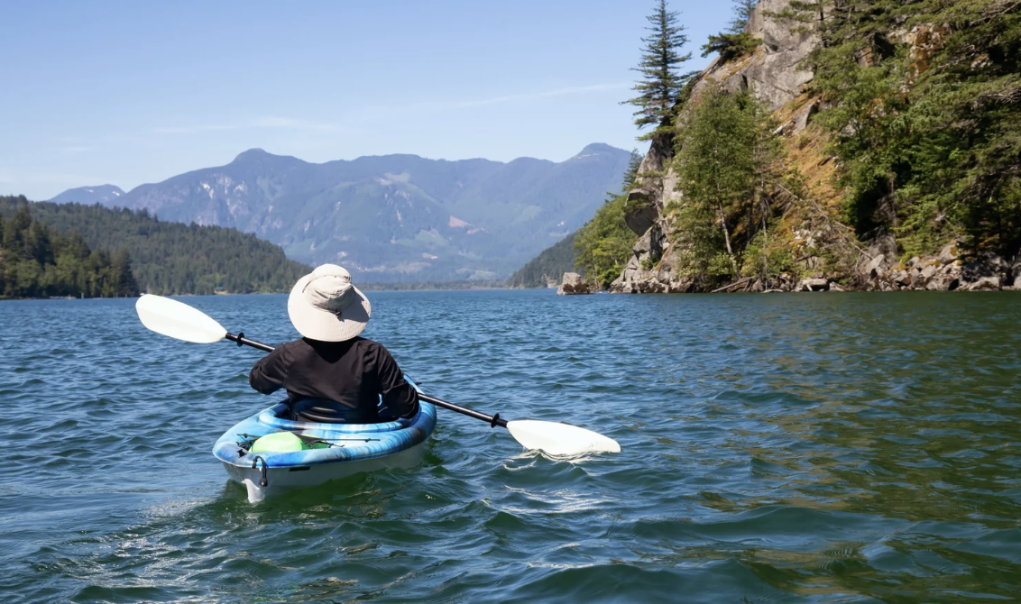 Person kayaking in a blue inflatable kayak on a lake surrounded by mountains and trees, with a clear blue sky.