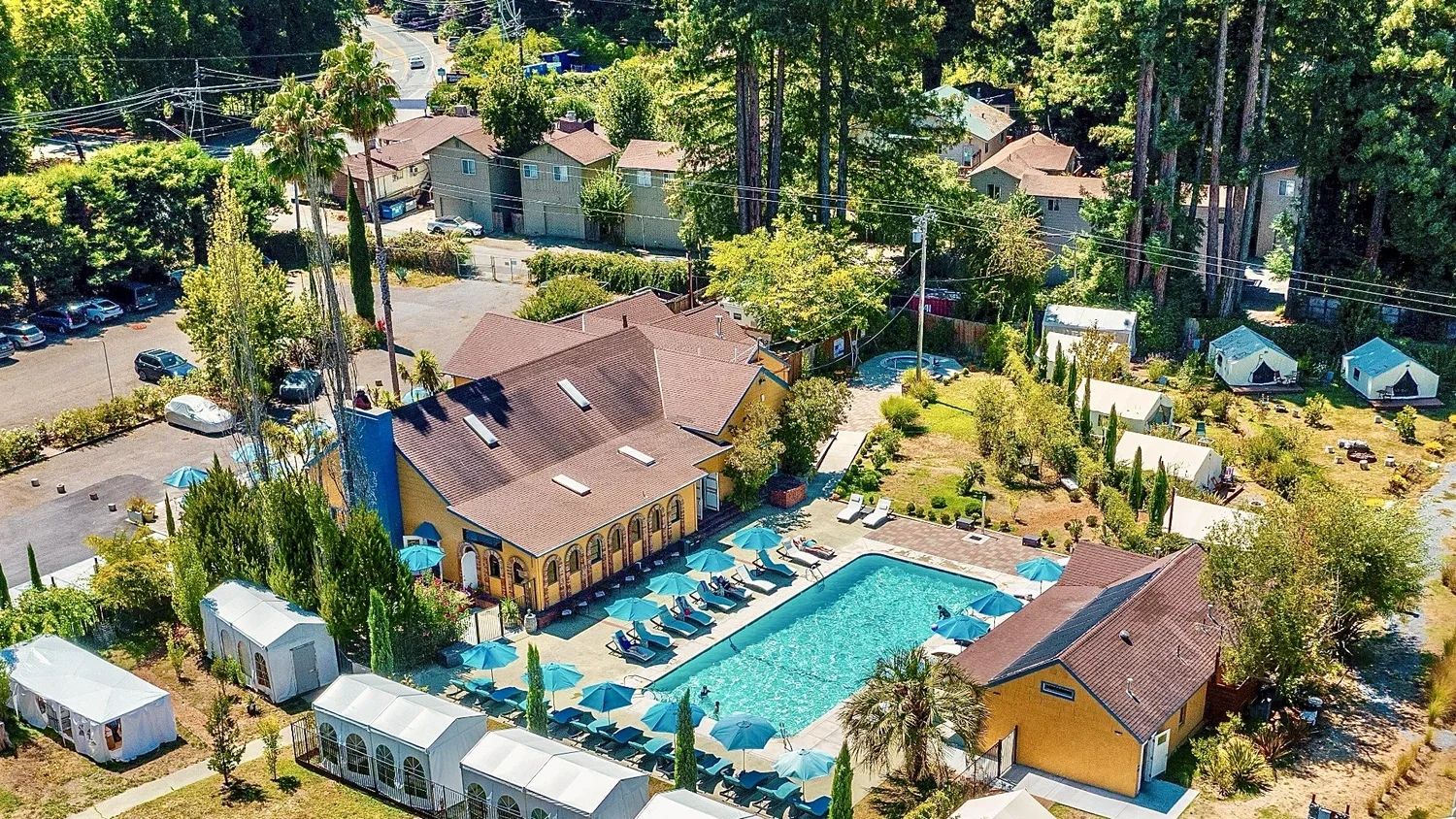 Aerial view of a residential outdoor swimming pool area with lounge chairs and umbrellas, surrounded by yellow buildings, trees, and nearby houses and parking lot.