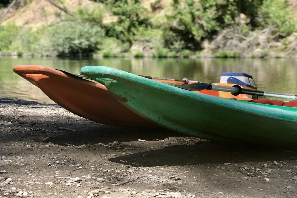 Close-up of a green and orange kayak resting on a dirt shore near a water body with trees in the background.