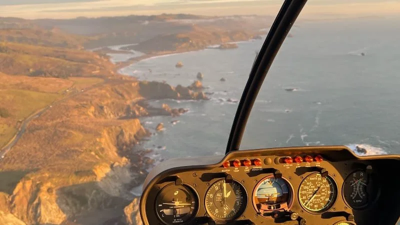 Aerial view of rugged coastline with cliffs and ocean waves seen from inside a helicopter cockpit.