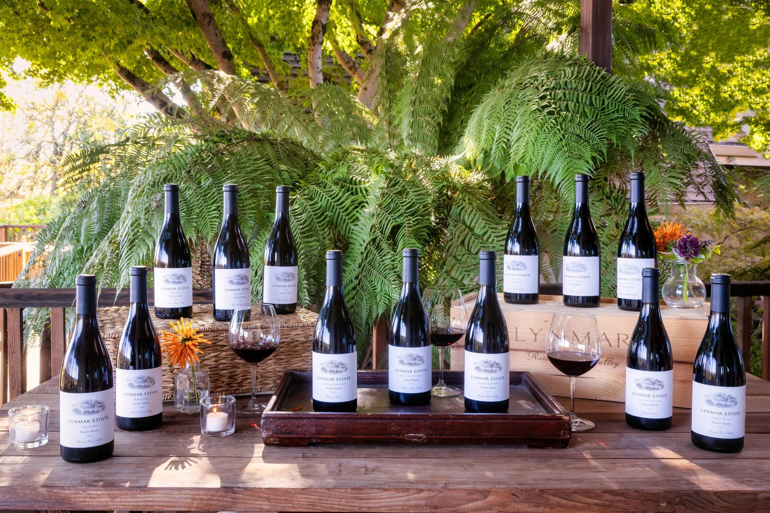 Multiple bottles of Lynmar Estate Pinot Noir wine on a rustic outdoor table with wine glasses, small candles, and a flower arrangement, set against lush green ferns and trees.