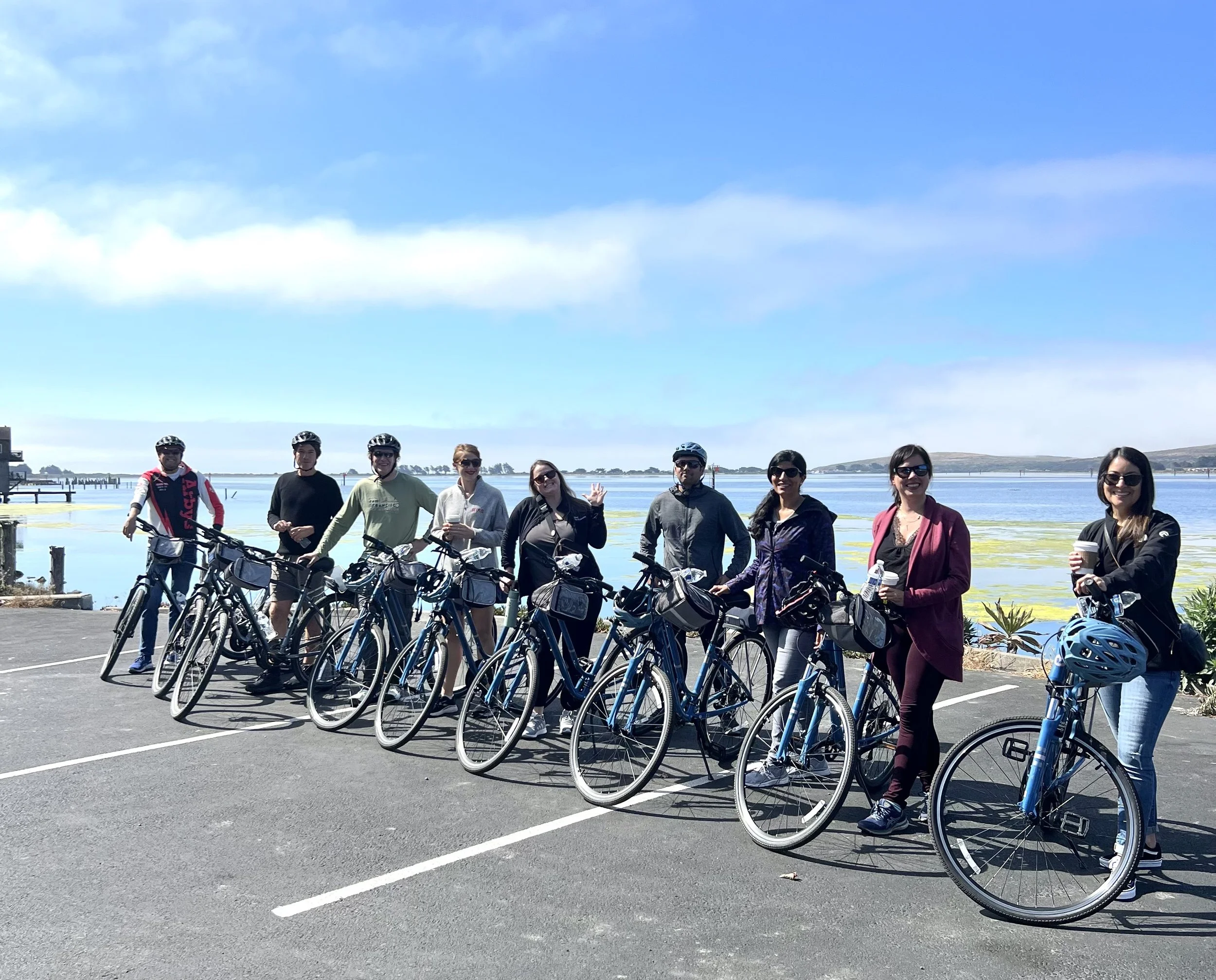 Group of ten people with bicycles near water and a pier under a partly cloudy sky.