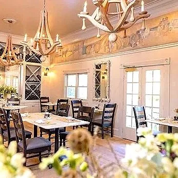 Elegant dining room with white walls, large windows, wooden chairs, and chandeliers, decorated with floral arrangements.