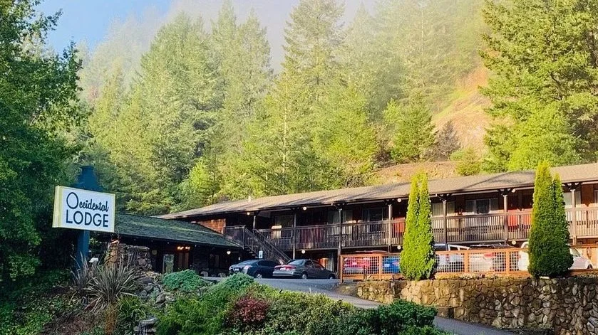 A two-story wooden lodge named 'Occidental LODGE' with parked cars in front, surrounded by green trees and a stone retaining wall.