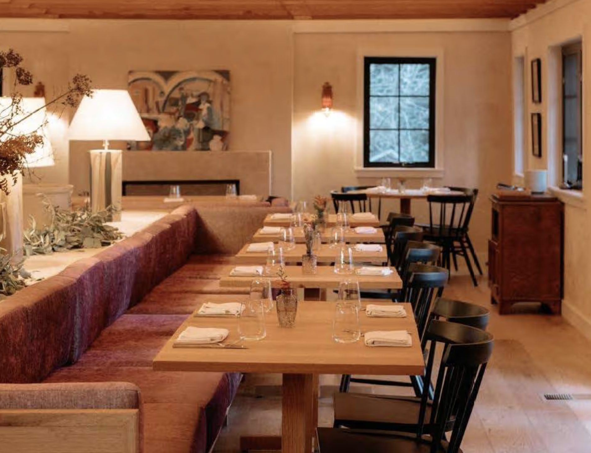 Empty dining room with wooden tables set with napkins, glasses, and vases with flowers. Black chairs surround the tables. Warm lighting from table lamps and wall sconces. Windows showing trees outside. Artwork on the walls.