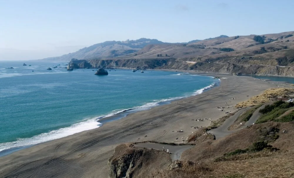 View of a rocky coastline with waves hitting the shore, cliffs in the distance, and a few boats offshore on a mostly clear day.