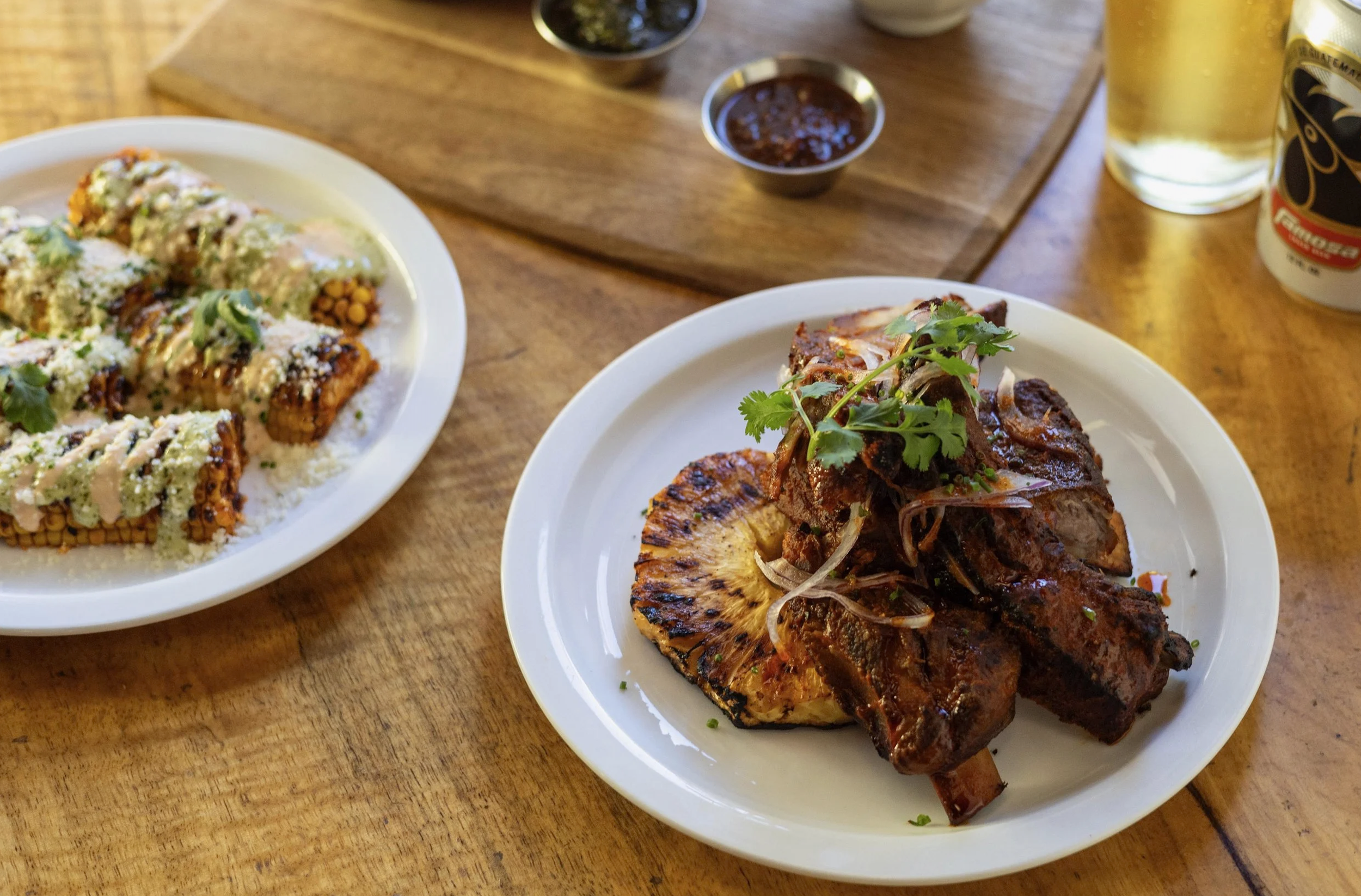 Close-up of plates of Mexican food on a wooden table, including grilled meat with pineapple slices, and a plate of sushi rolls with sauce and garnishes. There are small bowls of dipping sauces, a glass of beer, and a cup of coffee in the background.