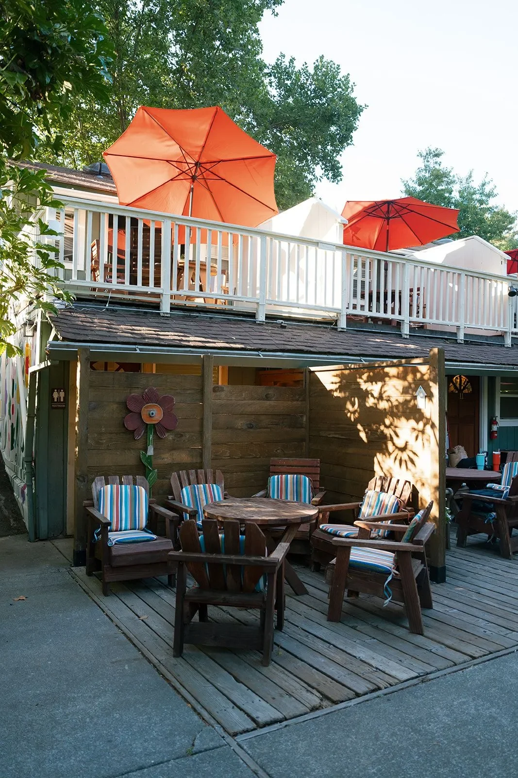 Outdoor patio with wooden chairs and tables, decorative flower wall art, and large orange and white umbrellas on the upper deck, with trees in the background.