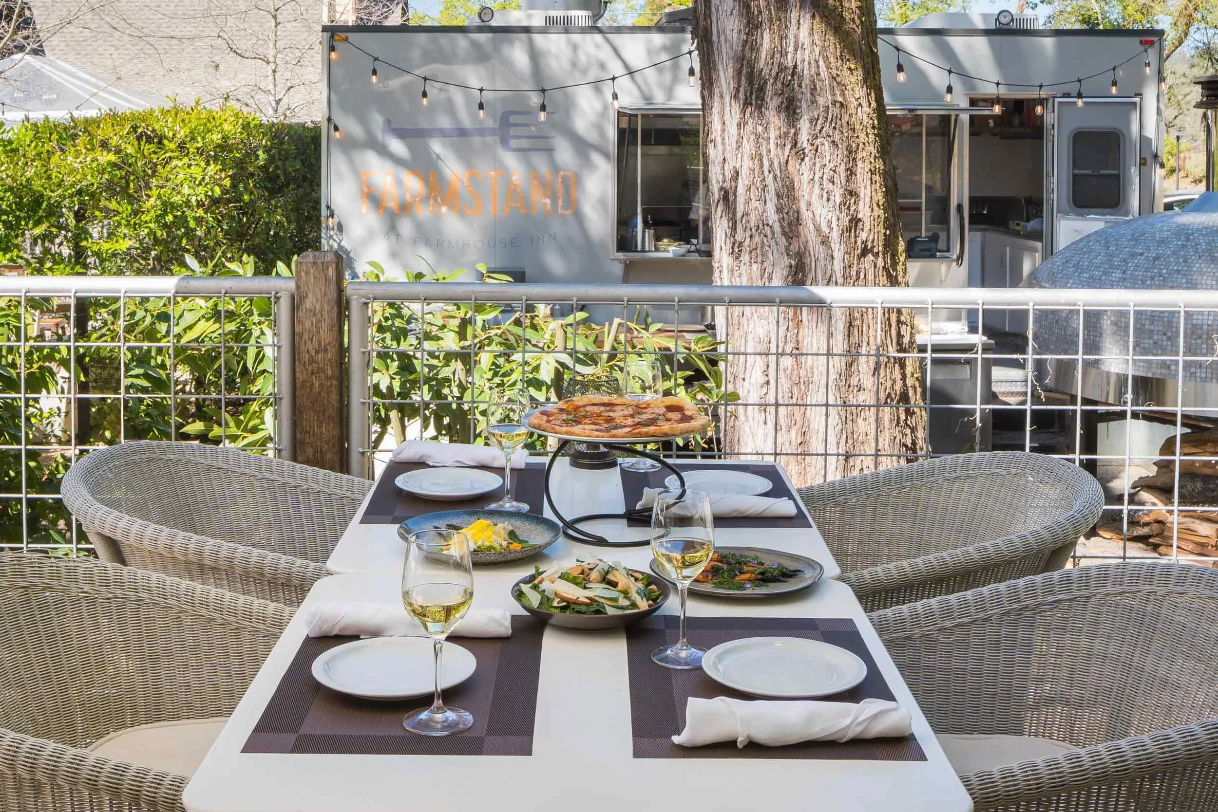 Outdoor dining table set with four glasses of white wine, plates, napkins, a pizza on a tray, and salads, with a food truck and trees in the background.