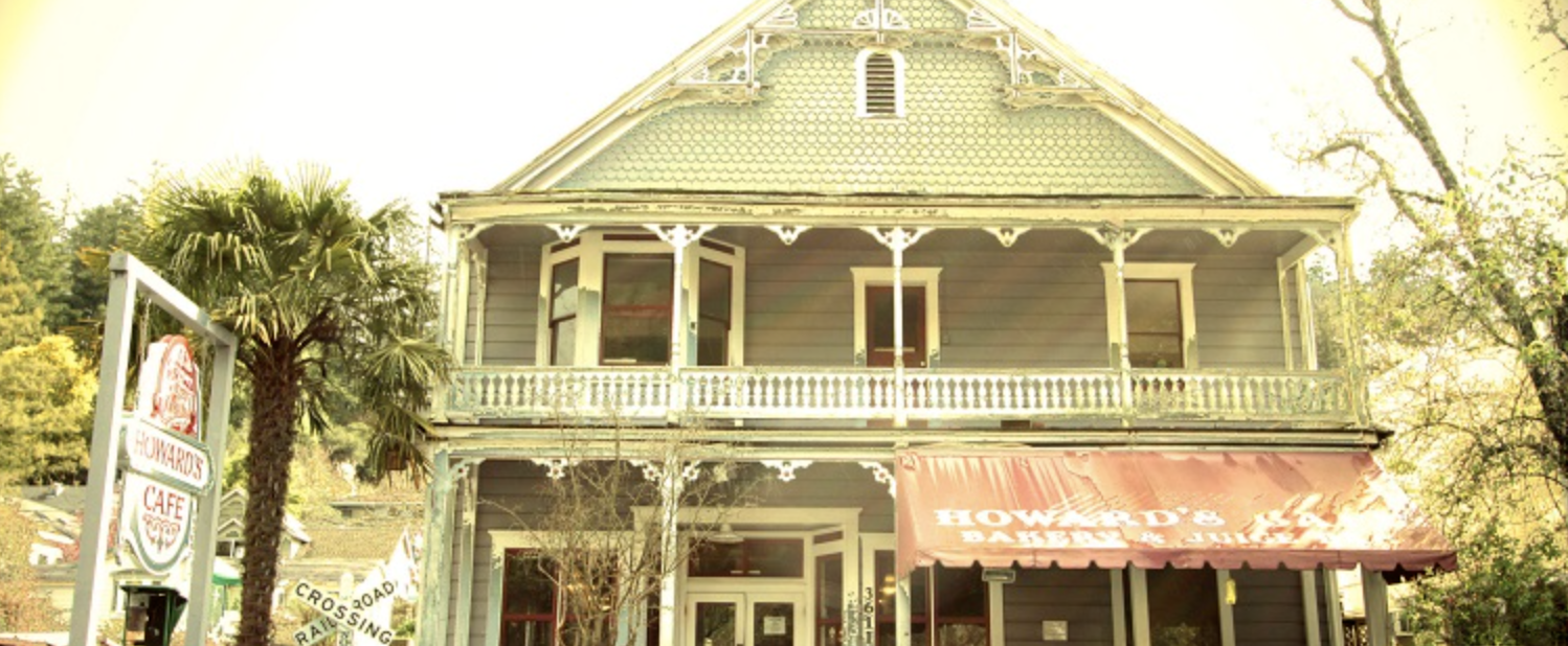 A two-story building with a greenish-gray exterior, a greenish roof, and a red awning that reads "HOWARD'S BAKE SHOP". There are open windows, a balcony on the second floor, and a sign on the left that says "HOWARD'S CAFE." Trees and other greenery surround the building.