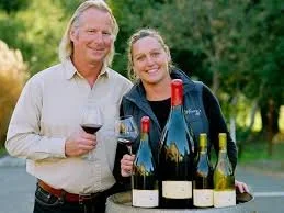 Two people, a man and a woman, smiling and holding glasses of red wine outdoors with a table of wine bottles in front of them.