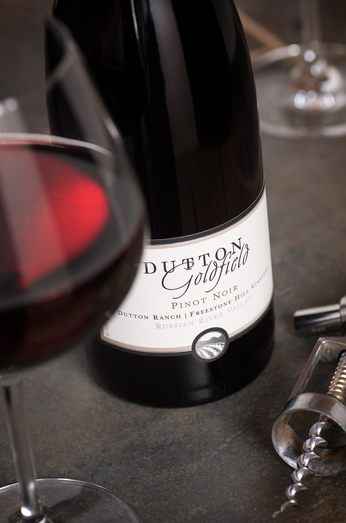 A bottle of Dutton Goldfield Pinot Noir wine next to a glass of red wine on a dark surface.