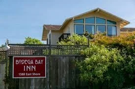 A house with large windows and a sloped roof, surrounded by greenery, located behind a wooden fence with a sign that reads 'Bodega Bay Inn, 1588 East Shore'.