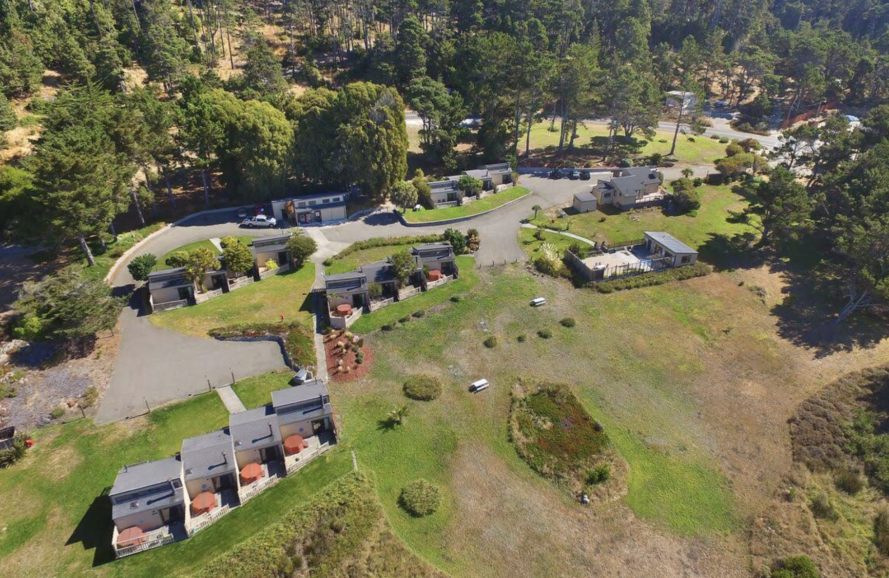 Aerial view of a residential area with small modern houses, green lawns, trees, and a winding driveway in a semi-rural setting with forested hills in the background.