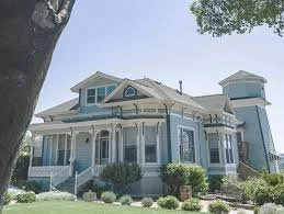 Large Victorian-style house with a wraparound porch and multiple turrets, surrounded by manicured bushes and trees.