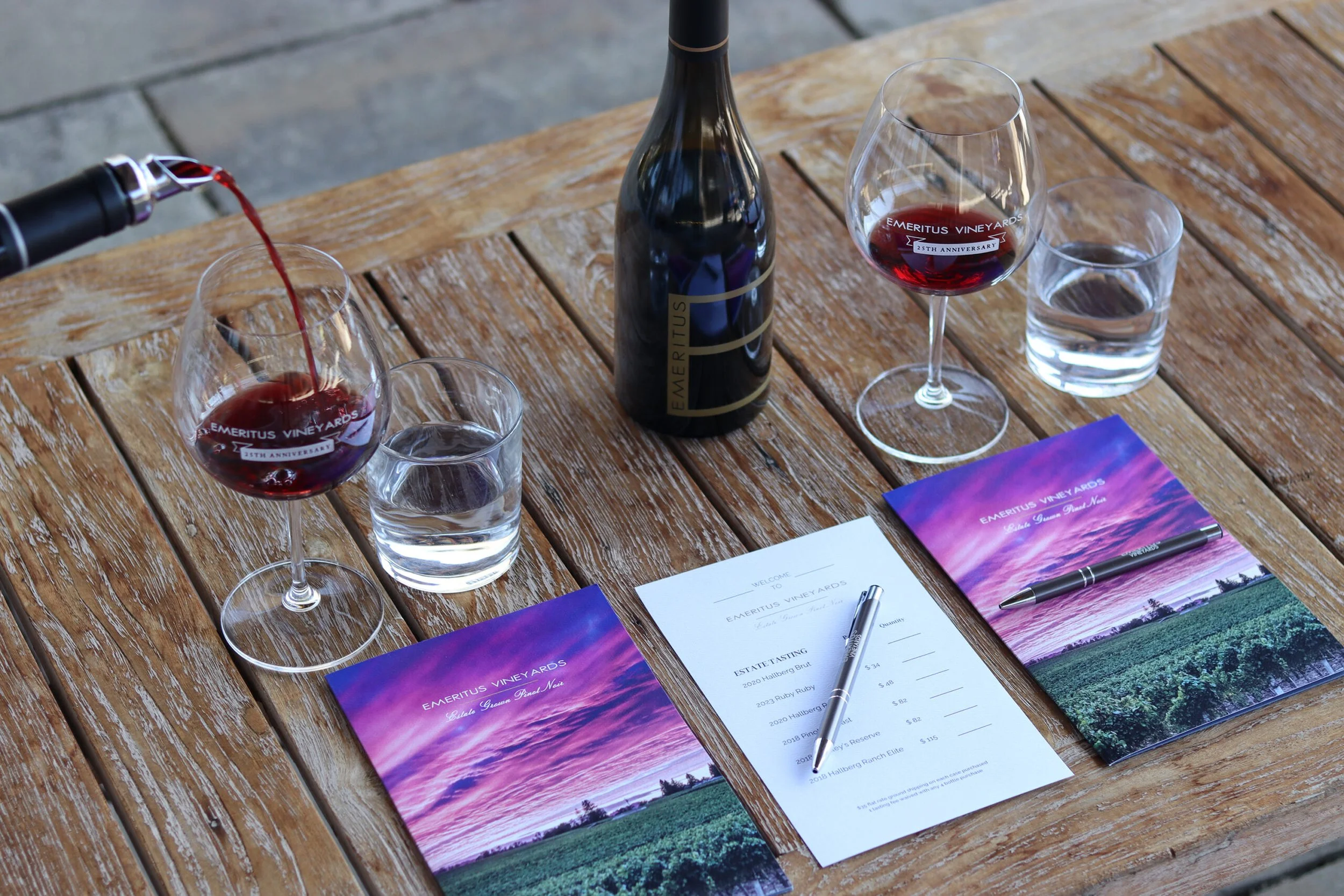 A wooden table with a bottle of Emeritus wine, two glasses of red wine, a glass of water, a list of estate tasting options, and two purple brochures with a sunset landscape design.