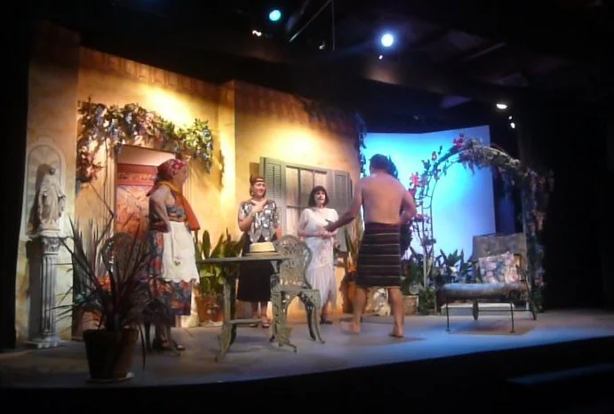 People performing a play on stage with a set designed to look like a garden or outdoor patio, decorated with flowers, plants, and outdoor furniture.