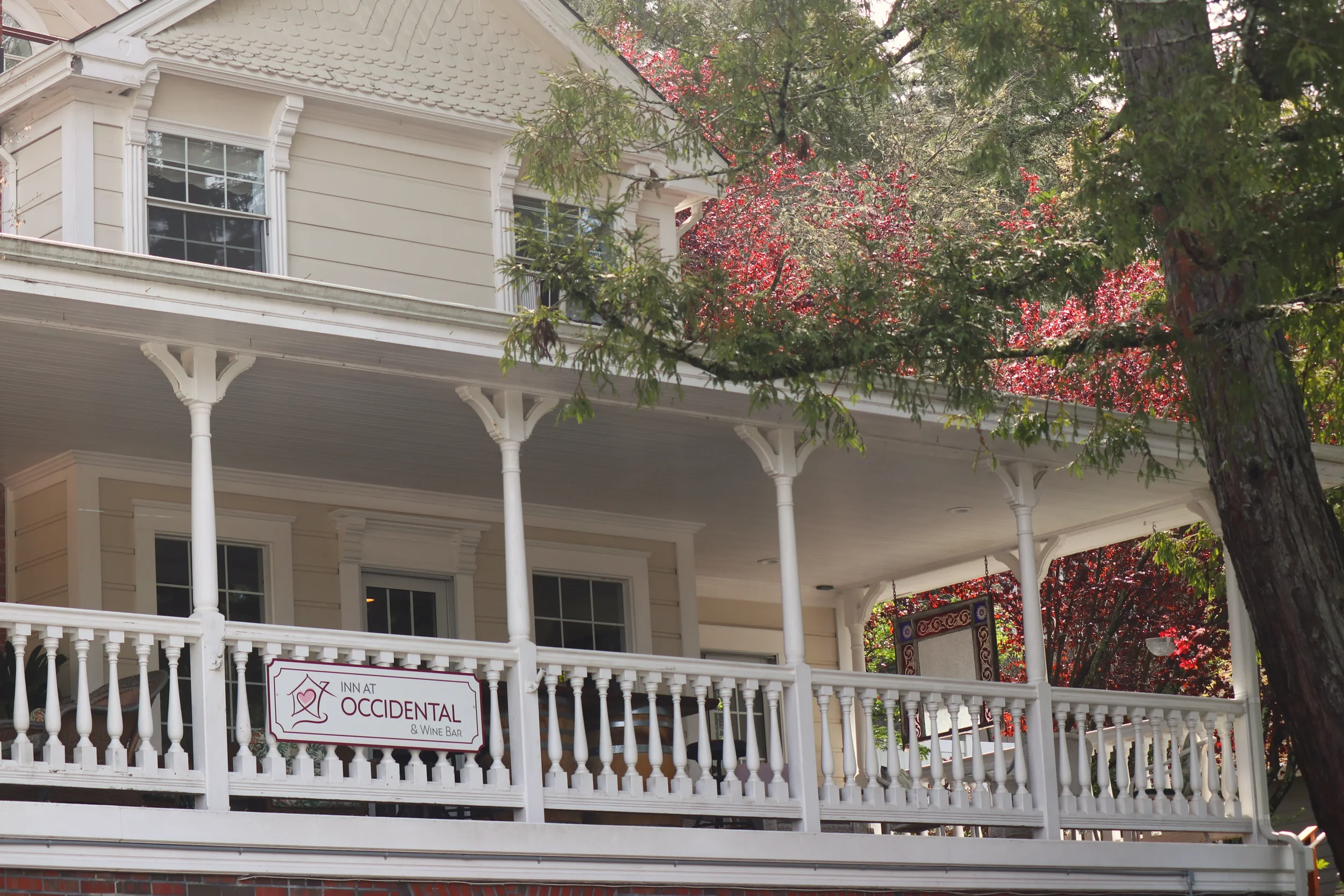 A cream-colored house with decorative trim and a porch, surrounded by green and red foliage. A sign on the porch reads 'Inn at Occidental & Wine Bar'.
