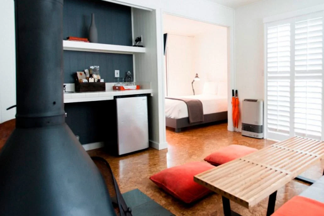 Living space with a black stove in the foreground, a wooden table with red cushions, a kitchenette with a mini refrigerator, and a bedroom with a bed and white shutters in the background.