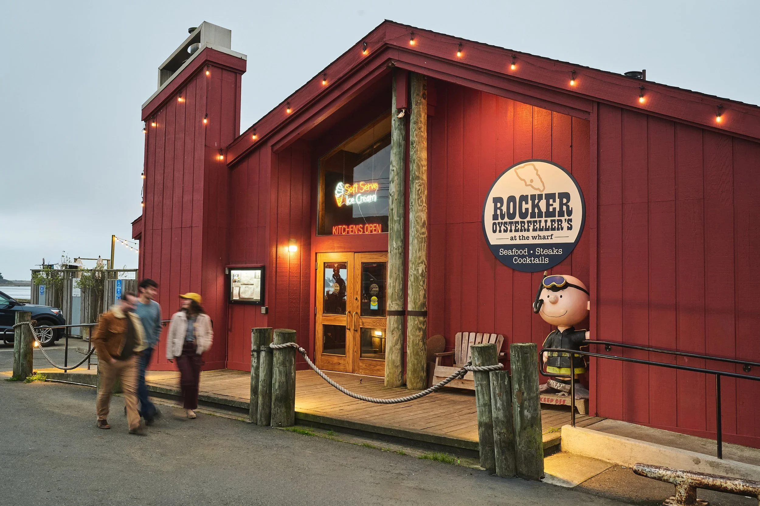 A red wooden restaurant building with string lights, a sign for Rocker Oysterfeller's, and a cartoon character statue near the entrance. There are three people walking past the restaurant at sunset.