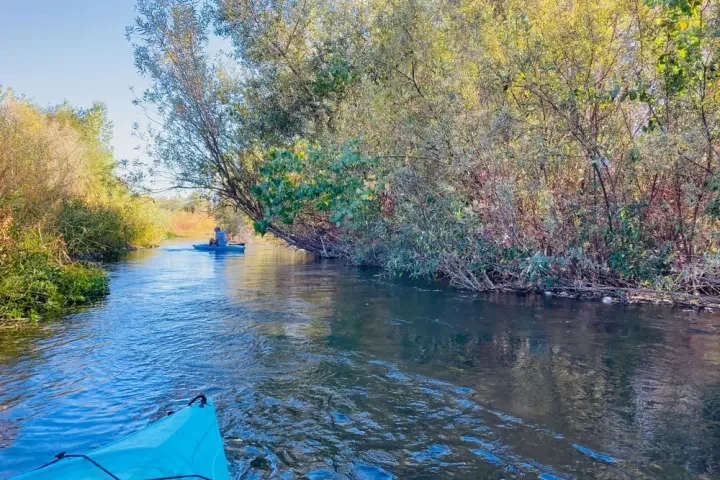 View of a peaceful waterway with a person kayaking in the distance and lush trees lining the water's edge on a sunny day.