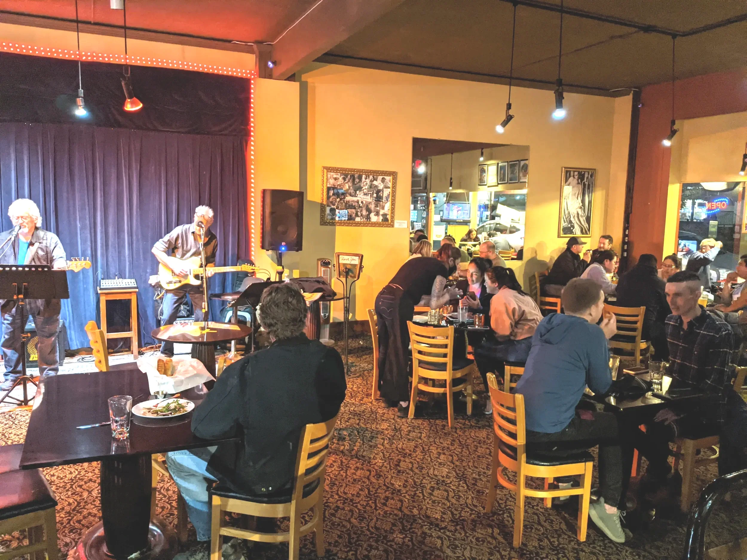 Live musical performance at a restaurant, with three musicians playing on a small stage, and diners seated at tables enjoying food and drinks in a cozy, vibrant atmosphere.