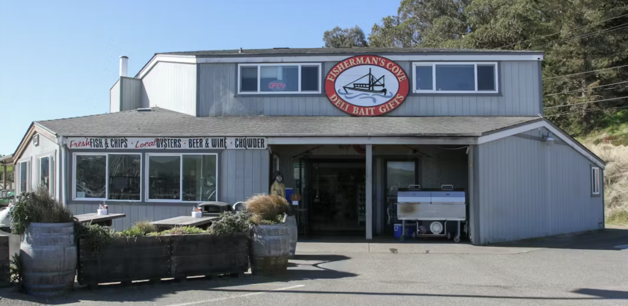 Exterior of Fishman's Cove, a seafood restaurant with a sign indicating fresh fish and chips, oysters, beer, wine, and chowder, featuring outdoor seating and a large logo with a fishing boat.