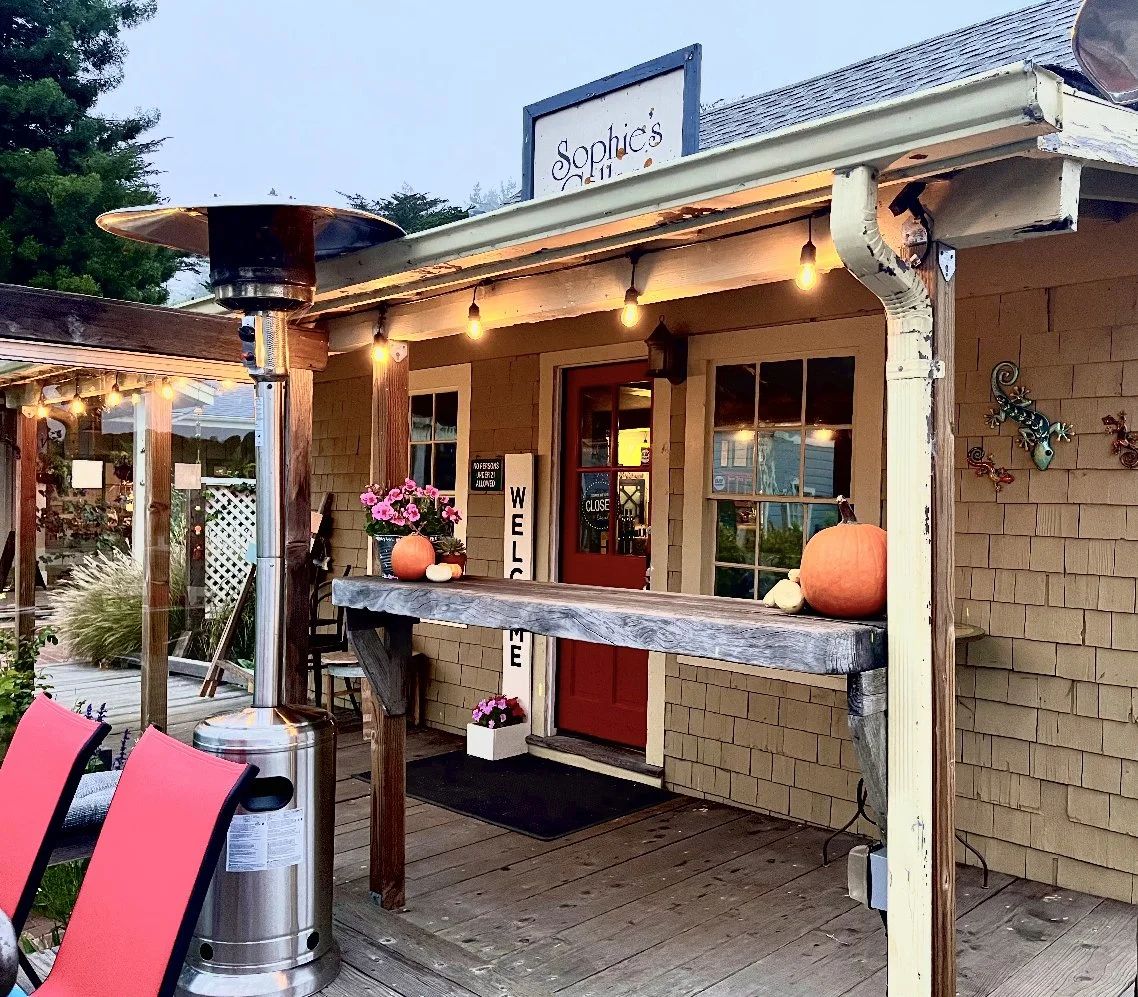Exterior of a cozy shop with string lights, autumn decorations, a red door, and outdoor seating.