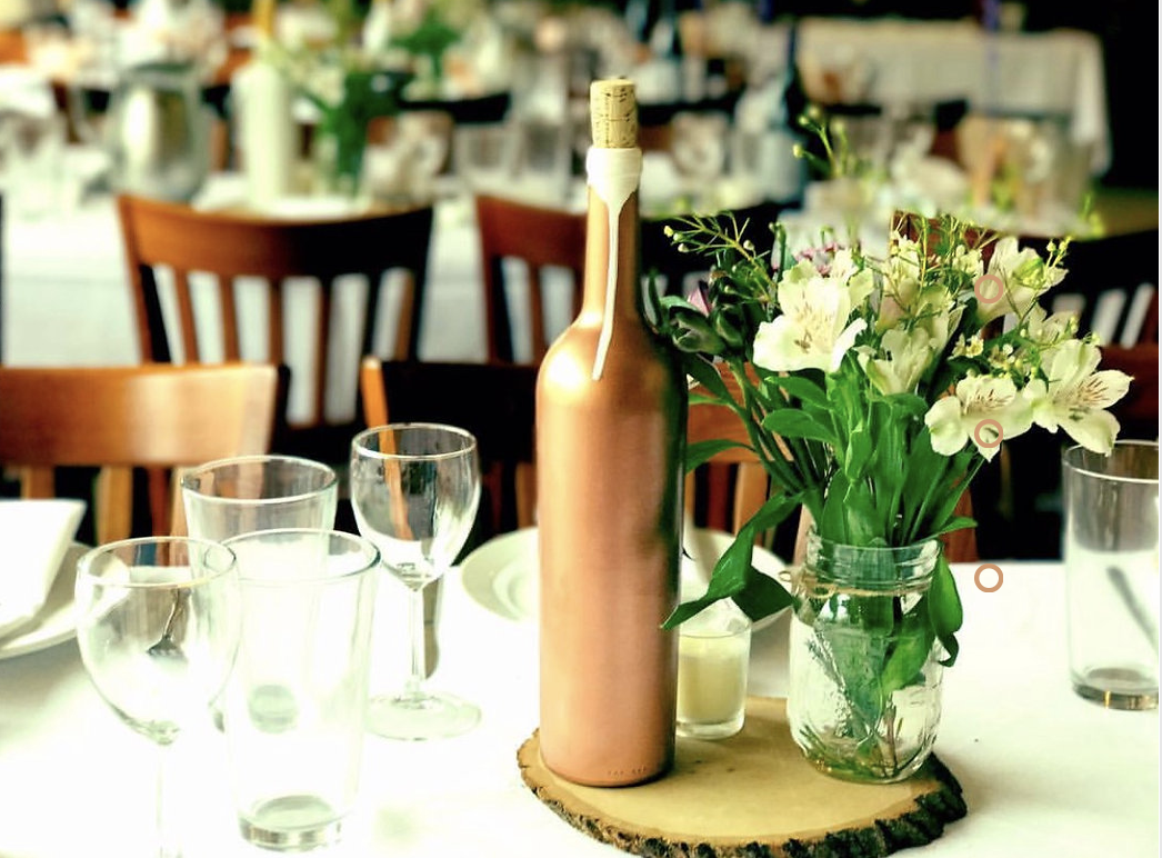 Table centerpiece with a pink wine bottle, a jar of white flowers, and several empty glasses, set in a restaurant with wooden chairs in the background.
