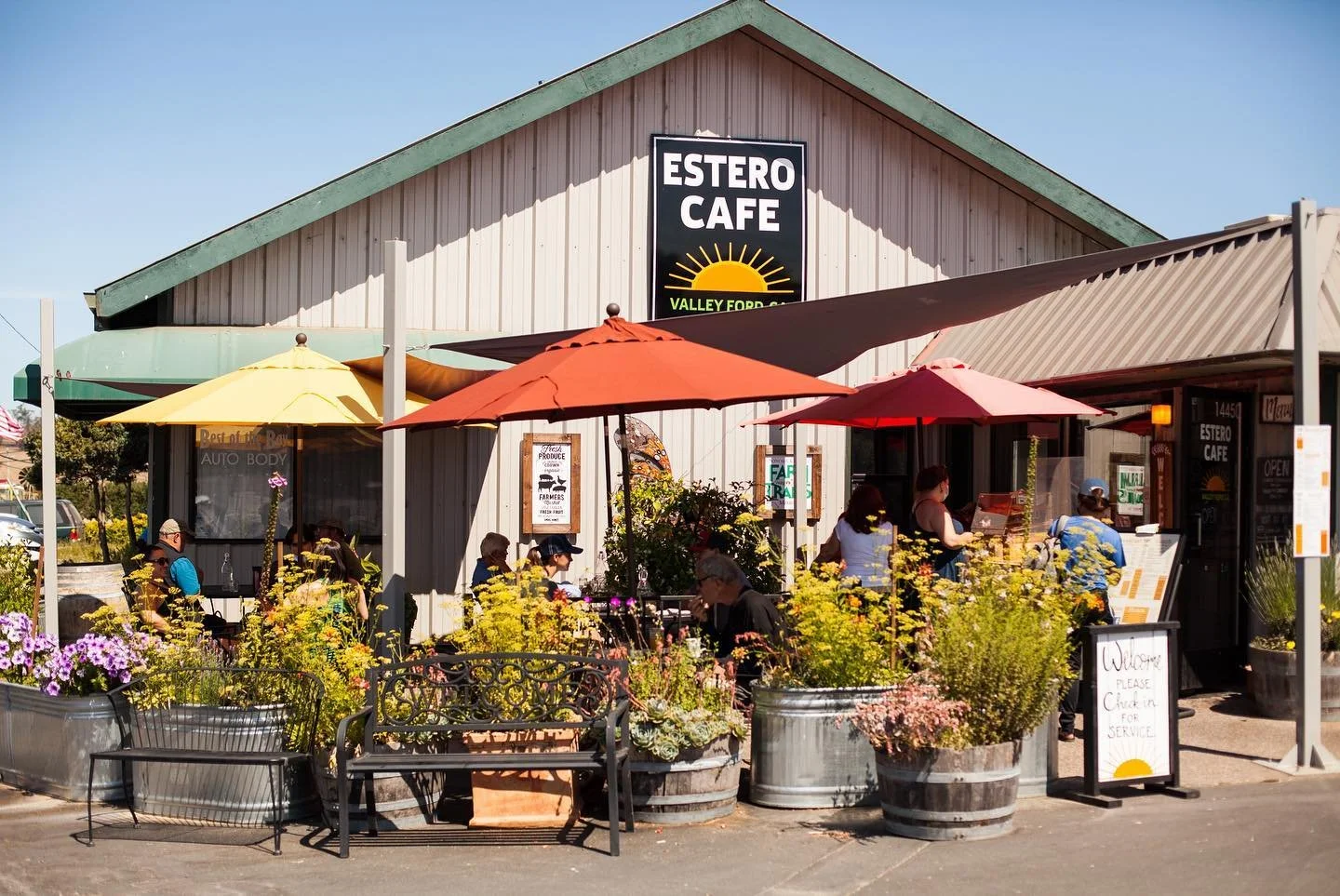 Outdoor seating area of Estero Cafe with colorful umbrellas, yellow, red, and brown, surrounded by potted plants and flowers, with people dining and a signboard at the entrance.