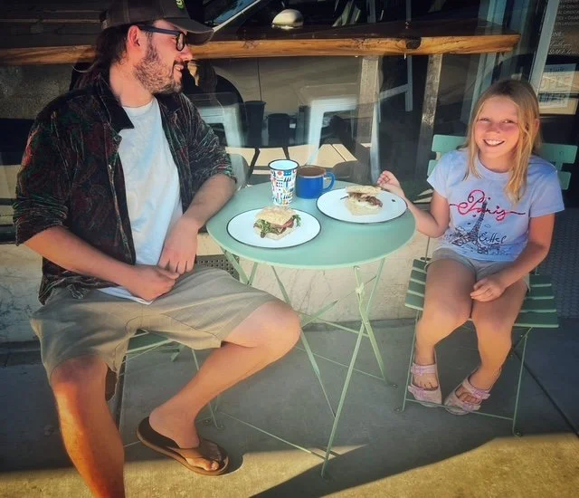 A man and a young girl sitting at a small outdoor table with plates of cake and cups, smiling and enjoying each other's company.