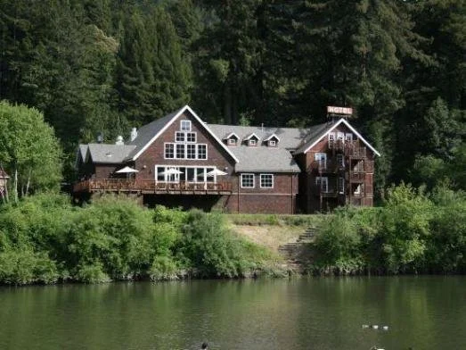 A large hotel with multiple levels and a balcony, situated by a body of water surrounded by dense trees.