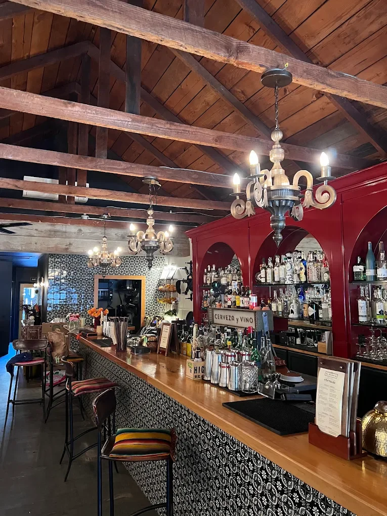 Inside a cozy bar with a wooden ceiling, vintage chandeliers, and a red bar counter stocked with bottles. There are bar stools with colorful cushions and decorative elements like flowers and framed signs.